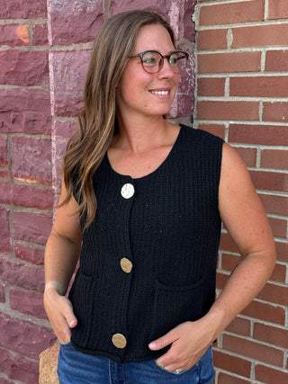 A lady wearing a black sweater vest with big gold buttons against a brick wall.