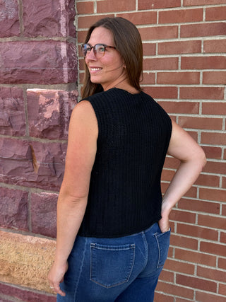 A lady wearing a black sweater vest with big gold buttons against a brick wall.
