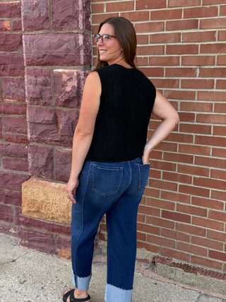 A lady wearing a black sweater vest with big gold buttons against a brick wall.
