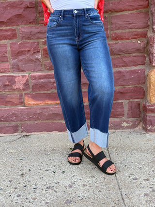 Lady wearing a barrel leg jean with a raw cuffed hem. Standing against a brick wall. 