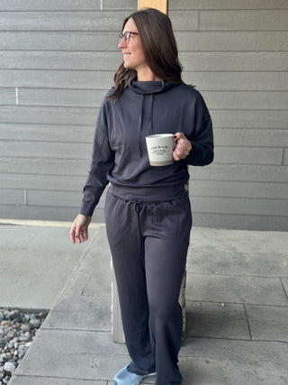Woman in dark gray hoodie and pants holding a coffee cup against a gray wall.