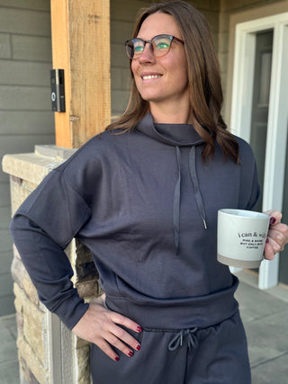 Woman in a dark hoodie holding a mug outside a house