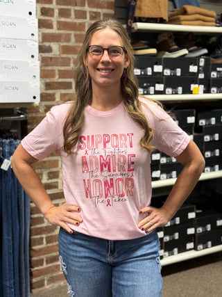A woman wearing a light pink graphic t-shirt with red text that reads 'SUPPORT THE FIGHTERS, ADMIRE THE SURVIORS, HONOR THE TAKEN and blue jeans, standing in a retail store with shelves of shoe boxes and clothing racks in the background.