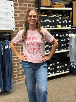 A woman wearing a light pink graphic t-shirt with red text that reads 'SUPPORT THE FIGHTERS, ADMIRE THE SURVIORS, HONOR THE TAKEN and blue jeans, standing in a retail store with shelves of shoe boxes and clothing racks in the background.