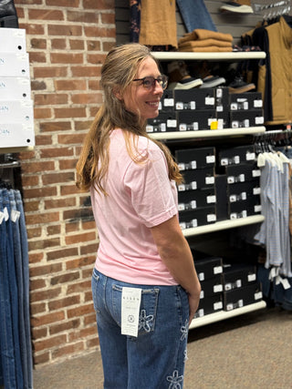 A woman with glasses and a ponytail stands in a clothing store, looking back over her shoulder. She is wearing a light pink t-shirt and blue jeans with decorative stitching on the back pocket.
