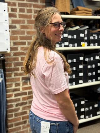 A woman with glasses and a ponytail, wearing a light pink t-shirt and blue jeans, standing in a retail store next to a brick wall with shelves of shoe boxes in the background.