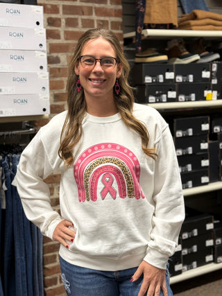 A woman wearing a white crewneck sweatshirt with a pink ribbon and leopard-print rainbow graphic, paired with blue jeans, standing in a retail store.