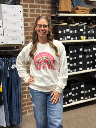 A woman wearing a white crewneck sweatshirt with a pink ribbon and leopard-print rainbow graphic, paired with blue jeans, standing in a retail store.