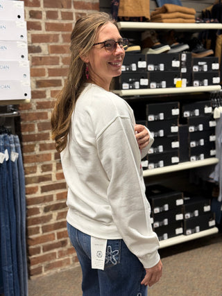 A woman wearing a cream-colored crewneck sweatshirt and blue jeans stands in a retail store, looking back over her shoulder. Shelves with black shoe boxes and a brick wall are visible in the background.