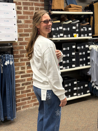A woman wearing a cream-colored crewneck sweatshirt and blue jeans stands in a retail store, looking back over her shoulder. Shelves with black shoe boxes and a brick wall are visible in the background.