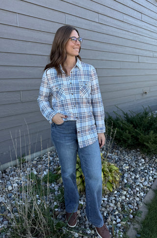 Woman wearing a plaid shirt and jeans standing in front of a gray wall.