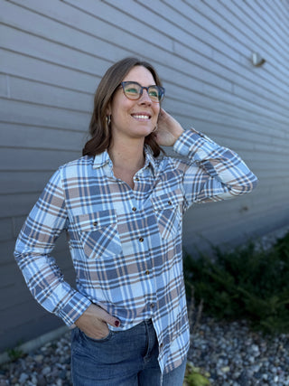 Woman wearing a plaid shirt and jeans standing against a gray wall.