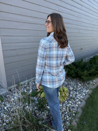 Woman wearing a plaid shirt and jeans standing against a gray wall.