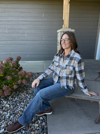 Woman sitting on a bench wearing a plaid shirt and jeans, with a gray wall and plants in the background.