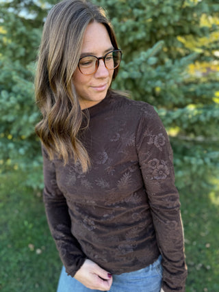 Woman wearing a brown floral-patterned sweater standing outdoors with greenery in the background