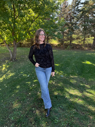Person wearing a black sweater and blue jeans standing in a grassy area with trees.