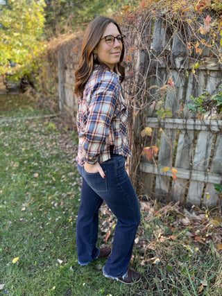 Woman wearing a plaid shirt and jeans standing in an outdoor setting with a wooden fence and autumn foliage.