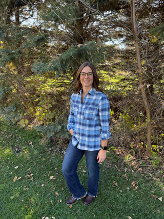 Person wearing a blue plaid shirt and jeans standing in a grassy area with trees.