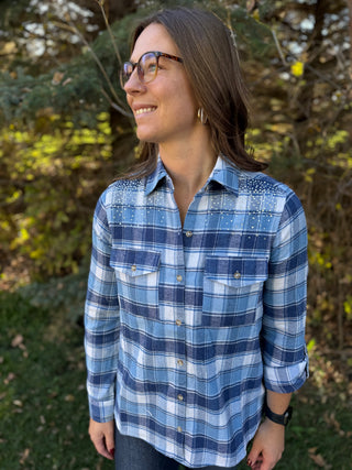 Woman wearing a blue plaid shirt standing outdoors with trees in the background