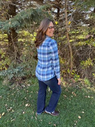 Woman wearing a blue plaid shirt and jeans standing in a grassy area with trees.