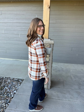 Woman wearing a plaid shirt and blue jeans standing on a concrete surface with a neutral wall in the background.