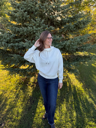 Woman in a white hoodie and blue jeans standing in a grassy area with trees.
