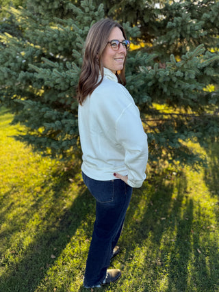 Woman wearing a white sweater and dark jeans standing outdoors with trees in the background