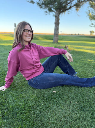 Woman sitting on grass wearing a pink shirt and blue jeans, with a tree and clear sky in the background.
