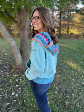 Woman wearing a light blue hoodie with a colorful pattern on the hood, standing outdoors in a park.