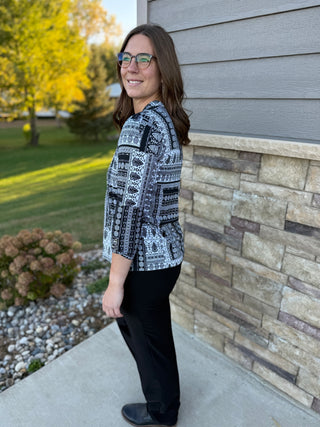 Woman standing outdoors next to a stone wall, wearing a patterned shirt and black pants.