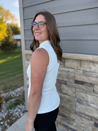Woman wearing a white sleeveless sweater against a brick wall.