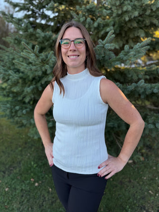 Woman wearing an ivory sleeveless sweater and dark pants standing outdoors with trees in the background.