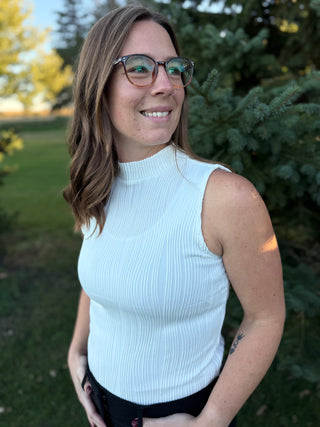 Woman wearing an ivory sleeveless top standing outdoors with trees in the background