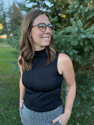 Woman wearing a black sleeveless top and gray pants standing outdoors with greenery in the background