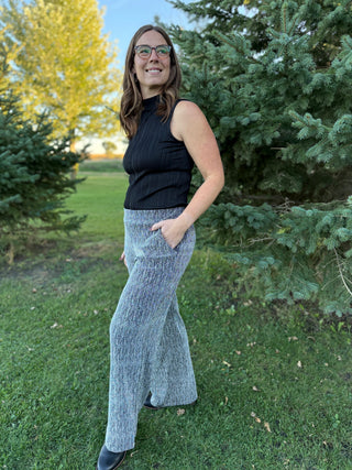 Woman in black sleeveless top and gray wide-leg pants standing outdoors with trees in the background