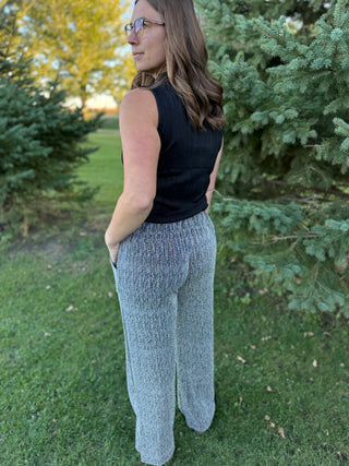 Woman wearing a black sleeveless top and light gray pants standing outdoors with trees in the background.