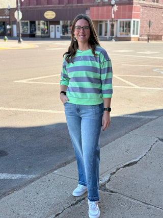 Woman wearing a green and purple striped sweater and blue jeans standing on a street corner.