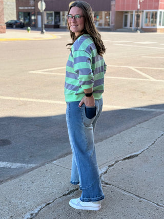 Woman wearing a striped sweater and jeans standing on a sidewalk with a parking lot in the background.