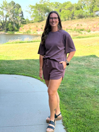 A woman wearing a matching mauve purple lounge set consisting of a short-sleeve t-shirt and drawstring shorts, standing outdoors on a paved path with a grassy park and a lake in the background.