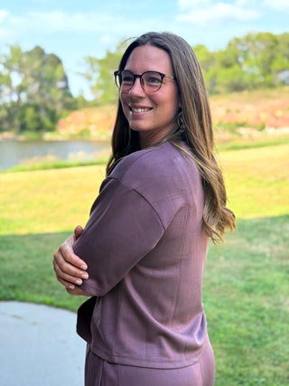 A woman with long brown hair and glasses, wearing a mauve-colored long-sleeve top and matching bottoms, standing outdoors on a golf course path with a blurred background of trees and grass.
