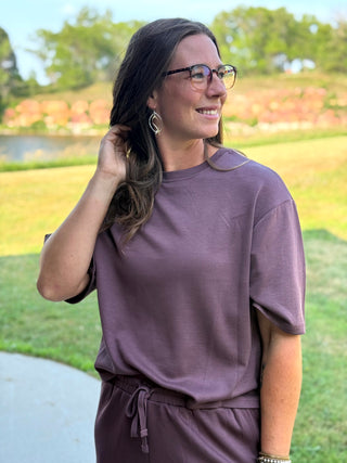 A woman wearing a matching mauve-purple lounge set consisting of a short-sleeve crewneck t-shirt and drawstring shorts, standing outdoors in a park setting.