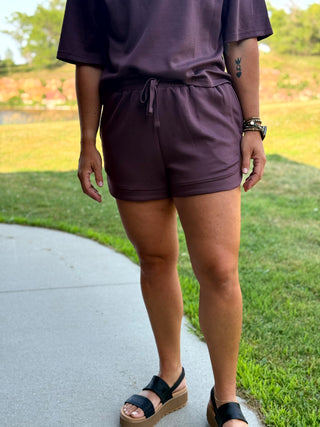 A person wearing a matching dark plum purple lounge set consisting of a short-sleeve top and drawstring shorts, standing outdoors on a concrete path next to a grassy lawn.