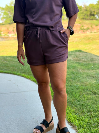 A person outdoors wearing a matching dark plum purple lounge set consisting of a short-sleeve top and drawstring shorts, paired with black platform sandals.