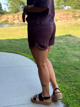 A person outdoors wearing a dark plum purple athletic set consisting of a short-sleeve top and matching shorts, paired with black platform sandals on a concrete path next to a grassy lawn.