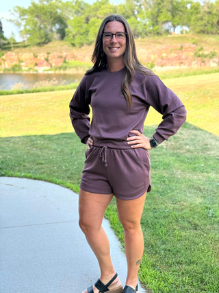 A woman wearing a matching mauve-brown lounge set consisting of a long-sleeve sweatshirt and drawstring shorts, standing outdoors on a paved path with a grassy park background.