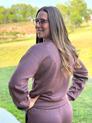 A woman with long brown hair and glasses, wearing a matching mauve-purple sweatshirt and shorts set, photographed from behind in an outdoor park setting.