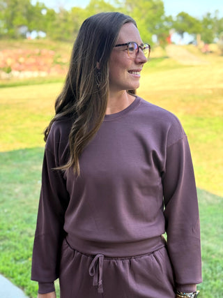 A woman wearing a matching mauve-brown sweatshirt and jogger pants set, standing outdoors on a grassy lawn.