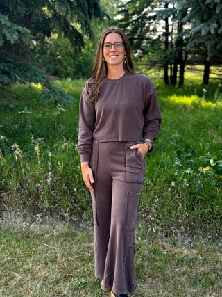 A woman wearing a matching two-piece lounge set in a dusty mauve-brown color stands outdoors in a grassy wooded area. The set includes a long-sleeve crewneck sweatshirt and matching high-waisted wide-leg pants with side pockets.