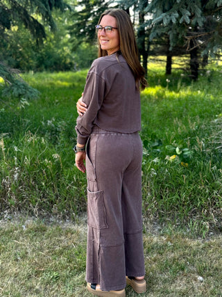 A woman wearing a matching two-piece lounge set in a dusty mauve-brown color stands outdoors in a grassy park area, viewed from behind.