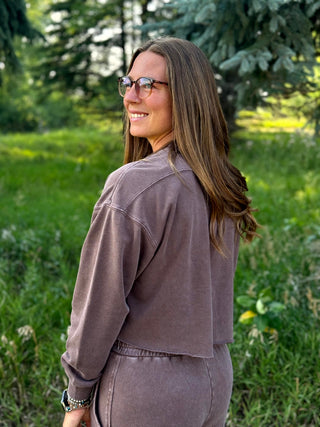 A woman with long brown hair and glasses, wearing a matching mauve-brown cropped sweatshirt and sweatpants set, standing outdoors in a grassy park area.